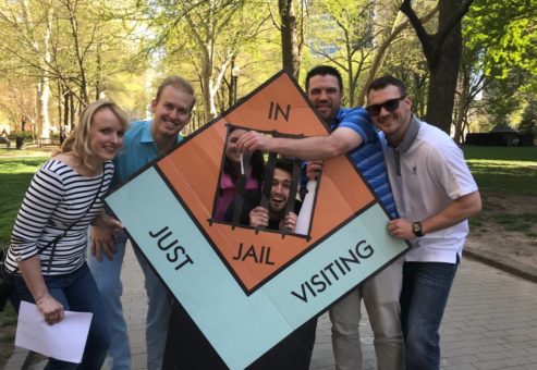 A group of six people smiling and posing with a large board game prop during a Team-opoly scavenger hunt, with one person playfully positioned behind the 'In Jail' section of the board.