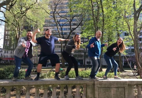 A team of five participants standing on a low stone wall, posing playfully during a Team-opoly scavenger hunt in a city park.