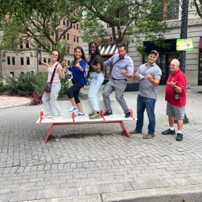 A group of team members standing in a fun pose on a public seesaw during a Team-opoly scavenger hunt team building event in an urban setting. thumbnail