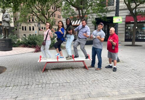 A group of team members standing in a fun pose on a public seesaw during a Team-opoly scavenger hunt team building event in an urban setting.
