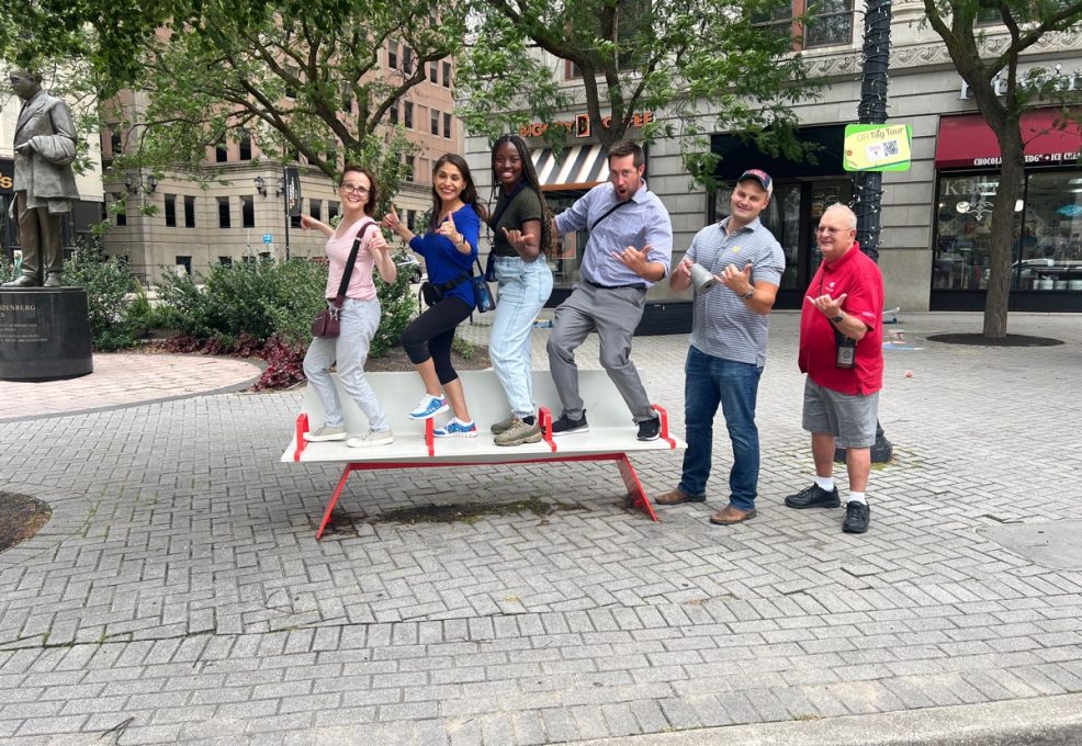 A group of team members standing in a fun pose on a public seesaw during a Team-opoly scavenger hunt team building event in an urban setting.