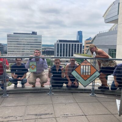 Team members posing behind a railing holding Monopoly-style 'In Jail' and 'Go to Jail' signs during a Team-opoly scavenger hunt team building event, with a cityscape in the background. thumbnail