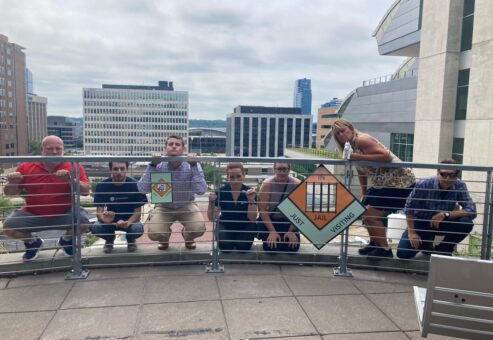 Team members posing behind a railing holding Monopoly-style 'In Jail' and 'Go to Jail' signs during a Team-opoly scavenger hunt team building event, with a cityscape in the background.