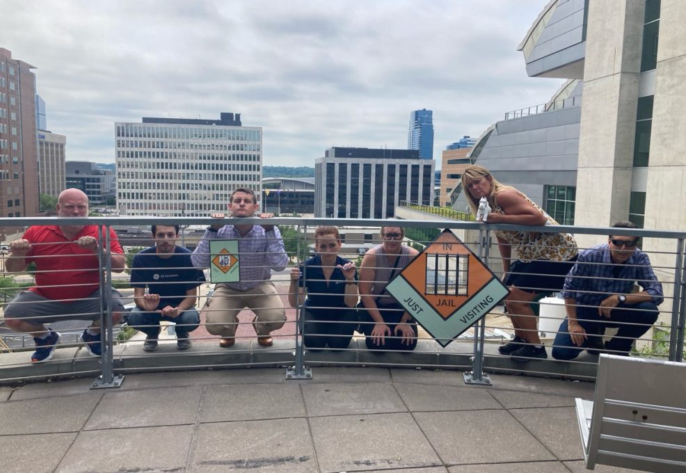 Team members posing behind a railing holding Monopoly-style 'In Jail' and 'Go to Jail' signs during a Team-opoly scavenger hunt team building event, with a cityscape in the background.