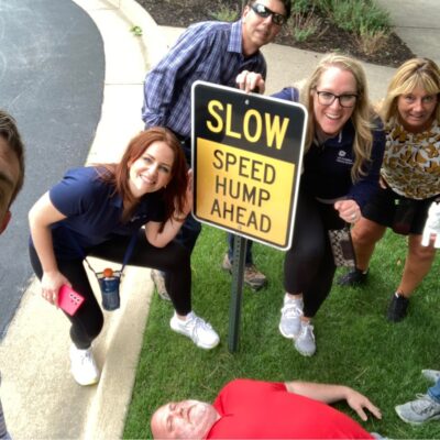 Team gathered around a 'Slow Speed Hump Ahead' sign, with one member humorously lying down next to it during a Team-opoly scavenger hunt team building event. thumbnail