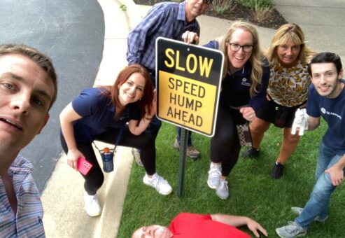 Team gathered around a 'Slow Speed Hump Ahead' sign, with one member humorously lying down next to it during a Team-opoly scavenger hunt team building event.