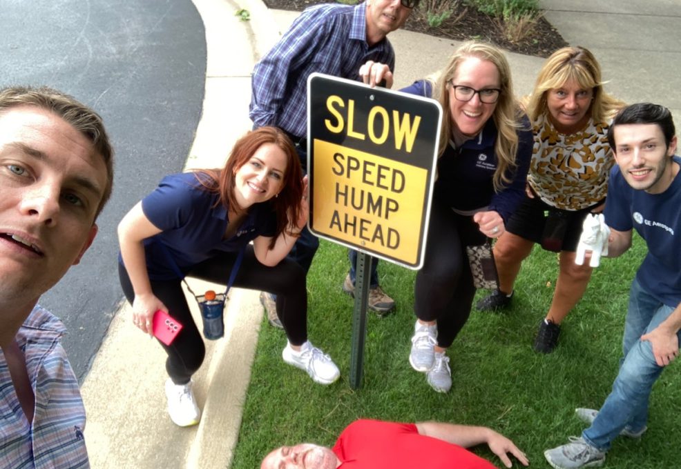 Team gathered around a 'Slow Speed Hump Ahead' sign, with one member humorously lying down next to it during a Team-opoly scavenger hunt team building event.