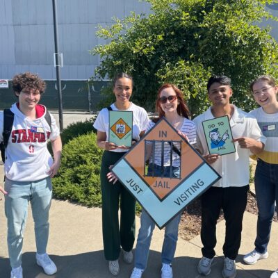 Group of participants posing with Monopoly-style 'In Jail' and 'Go to Jail' signs during a Team-opoly scavenger hunt outdoors, with smiles and playful expressions. thumbnail