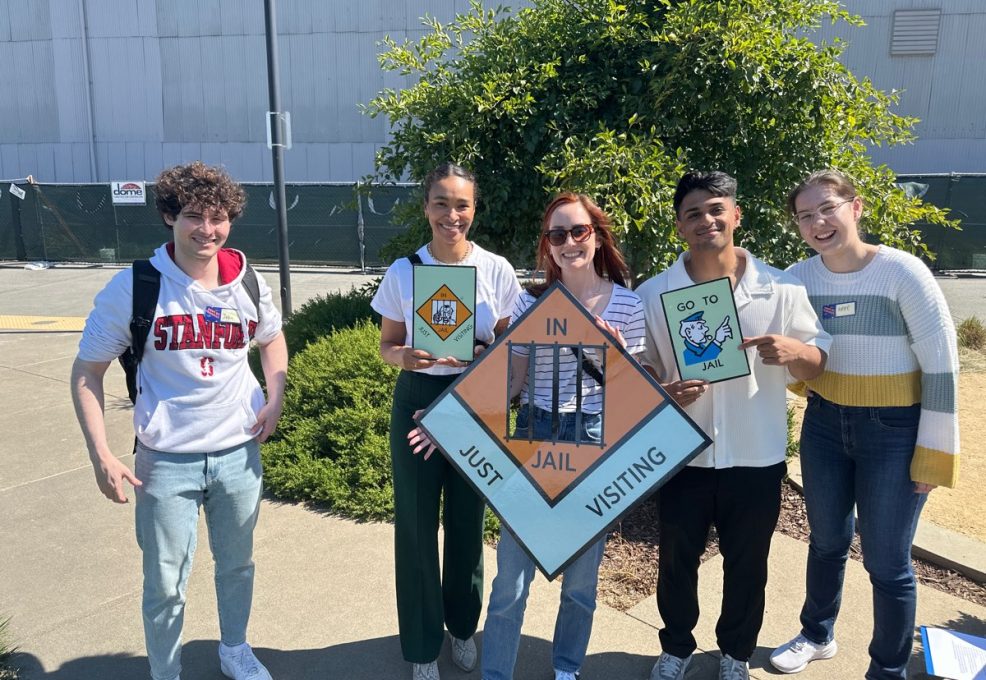 Group of participants posing with Monopoly-style 'In Jail' and 'Go to Jail' signs during a Team-opoly scavenger hunt outdoors, with smiles and playful expressions.