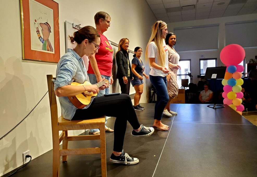 Participants perform on stage during a team building event. One person sits and plays the ukulele while others stand nearby, preparing to sing or dance as part of the Team Ukulele activity.