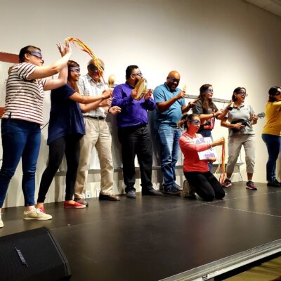 A group of participants stands on stage, wearing blindfolds and holding various props, including ukuleles and ribbons, as part of a team building event. They are engaged in a collaborative performance during the Team Ukulele activity. thumbnail