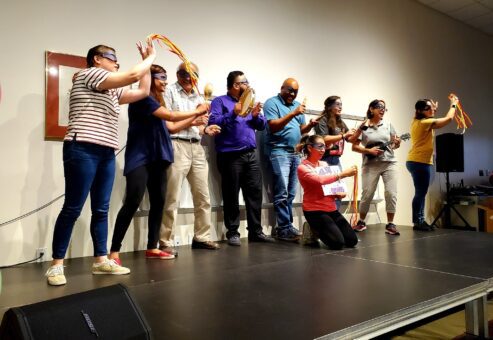 A group of participants stands on stage, wearing blindfolds and holding various props, including ukuleles and ribbons, as part of a team building event. They are engaged in a collaborative performance during the Team Ukulele activity.