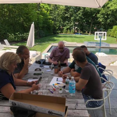 Participants sit around an outdoor table in a backyard setting, brainstorming and preparing for a team building activity as part of the Team Ukulele event. The group is focused, with materials like markers and ukulele parts spread across the table. thumbnail