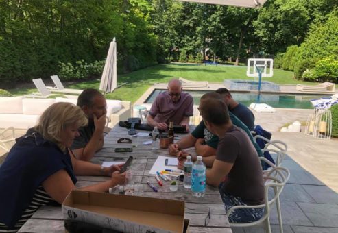 Participants sit around an outdoor table in a backyard setting, brainstorming and preparing for a team building activity as part of the Team Ukulele event. The group is focused, with materials like markers and ukulele parts spread across the table.