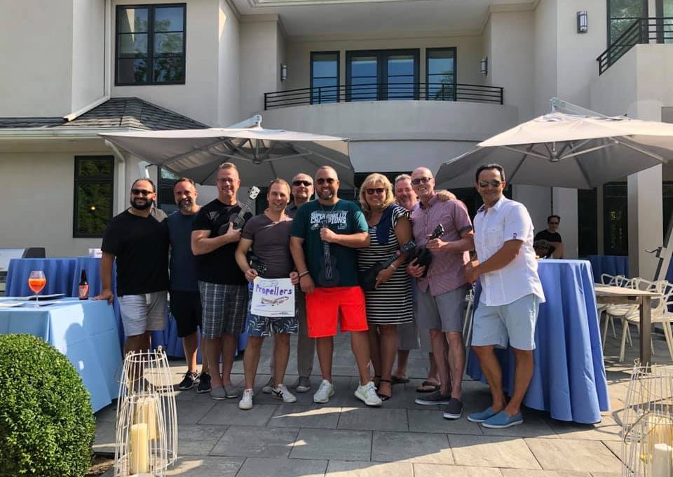 A group of participants stands together outdoors in front of a modern building, smiling and posing for a photo during a team building event. Some hold ukuleles.