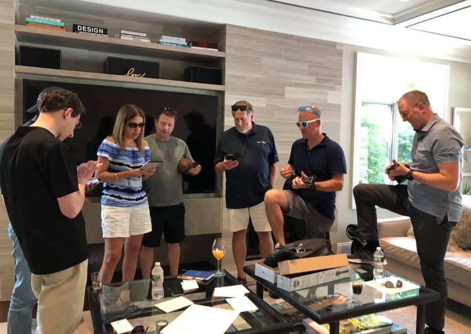 A group of participants stands in a casual living room setting, each holding a ukulele and focusing on learning chords during a team building event. Boxes and papers are scattered on the table as part of the Team Ukulele activity.