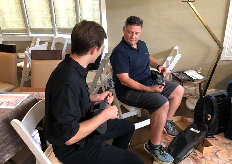 Two participants sitting and holding ukuleles during a team building event. They are engaged in conversation, learning to play their instruments as part of the Team Ukulele music activity.
