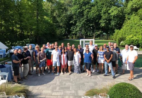 A large group of participants gathers for a group photo outdoors in a scenic garden area during a team building event. The group stands together smiling, enjoying the Team Ukulele activity.