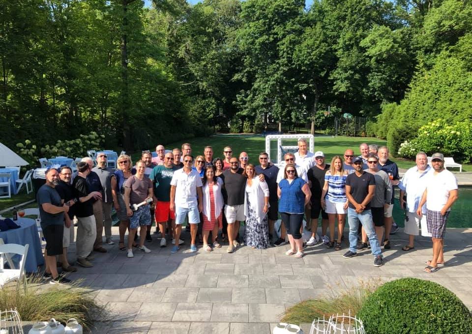 A large group of participants gathers for a group photo outdoors in a scenic garden area during a team building event. The group stands together smiling, enjoying the Team Ukulele activity.