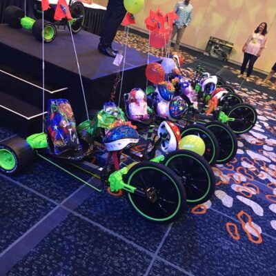 A row of assembled green Green Machine bikes is displayed on the floor, each adorned with colorful decorations, helmets, and balloons as part of a team building event. thumbnail