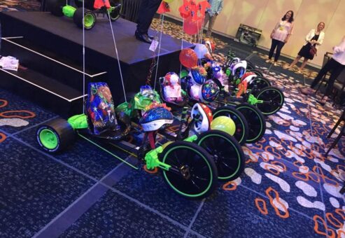 A row of assembled green Green Machine bikes is displayed on the floor, each adorned with colorful decorations, helmets, and balloons as part of a team building event.