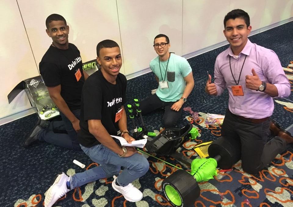 Four individuals kneeling on the floor assembling a Green Machine bike during a team building event. One person gives a thumbs-up while others smile, holding tools and instructions, as they work together on the project.
