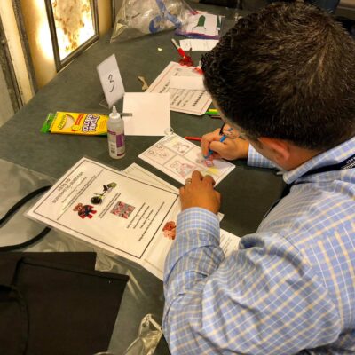 A man seated at a table is focused on drawing and coloring in a comic-style booklet. Various sheets of paper with superhero-themed drawings and instructions are spread out in front of him, along with a pack of colored markers and a bottle of glue. The scene captures creativity and concentration, as he works on a charitable team building activity. thumbnail