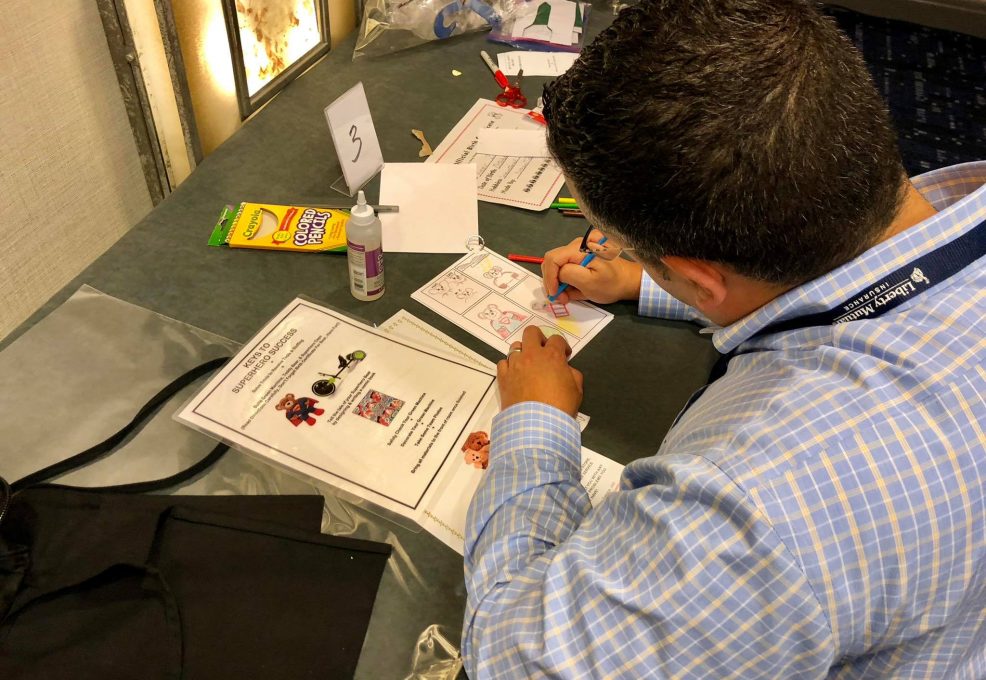A man seated at a table is focused on drawing and coloring in a comic-style booklet. Various sheets of paper with superhero-themed drawings and instructions are spread out in front of him, along with a pack of colored markers and a bottle of glue. The scene captures creativity and concentration, as he works on a charitable team building activity.