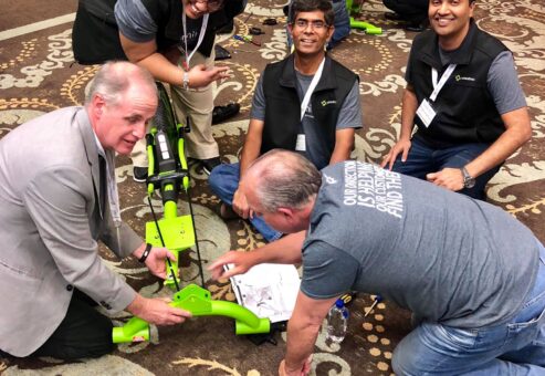 A group of five people, some seated and others kneeling, work together to assemble a bright green bike. Two large wheels are placed on the floor next to the bike frame. The team appears to be engaged and smiling, collaborating on the project in a room filled with other teams working on similar bikes as part of a charitable team building activity.