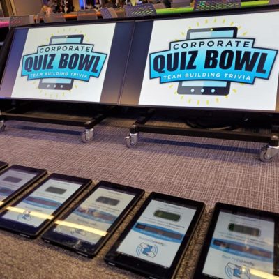 Corporate Quiz Bowl setup featuring large screens displaying the logo and a row of tablets ready for team participation in this engaging team building trivia event.