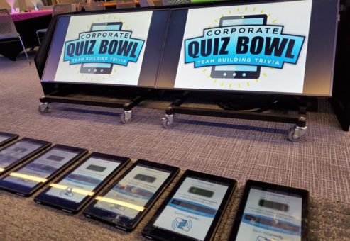 Corporate Quiz Bowl setup featuring large screens displaying the logo and a row of tablets ready for team participation in this engaging team building trivia event.