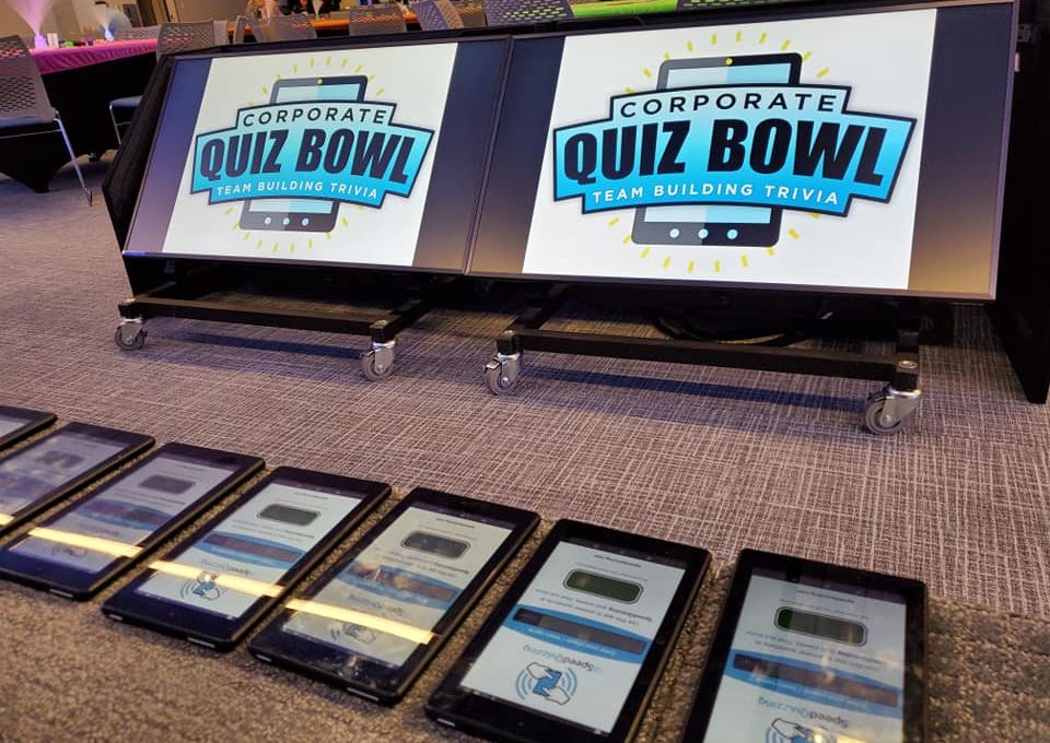 Corporate Quiz Bowl setup featuring large screens displaying the logo and a row of tablets ready for team participation in this engaging team building trivia event.