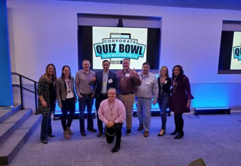 Smiling participants pose in front of a Corporate Quiz Bowl screen after completing a lively team building trivia competition.