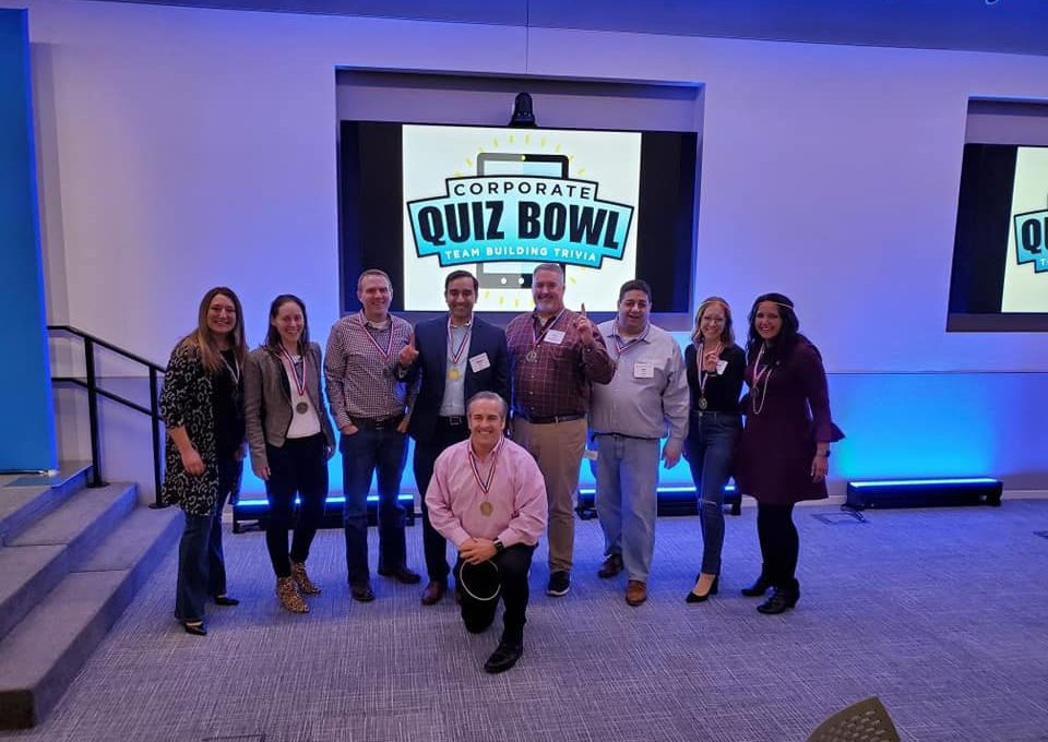 Smiling participants pose in front of a Corporate Quiz Bowl screen after completing a lively team building trivia competition.
