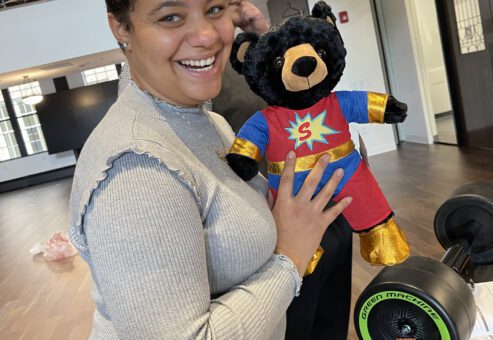 A smiling woman holding a black teddy bear dressed in a superhero outfit, complete with a red suit, gold accents, and a large 'S' emblem on the chest. In the background, a Green Machine bike with neon green wheels is visible.