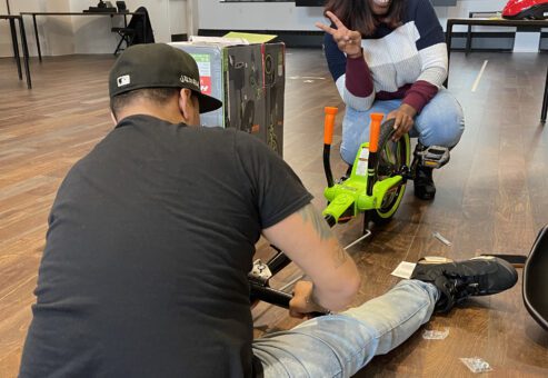 A man sitting on the floor assembling a Green Machine bike as part of a charitable team building event while a woman kneels behind it, flashing a peace sign and smiling.