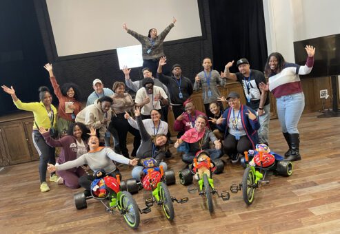 A group of people posing energetically with their arms raised, standing behind assembled Green Machine bikes. They are celebrating the accomplishment of a successful charitable team building event.