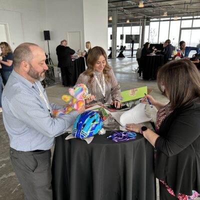 Three people stand around a high-top table, collaborating on assembling items. One person holds a colorful stuffed bear, while another is focused on a helmet and decorations. The third person writes notes on a document. They are participating in a charitable team building activity. thumbnail