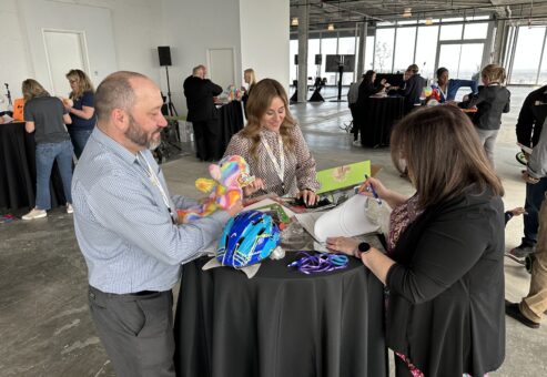 Three people stand around a high-top table, collaborating on assembling items. One person holds a colorful stuffed bear, while another is focused on a helmet and decorations. The third person writes notes on a document. They are participating in a charitable team building activity.