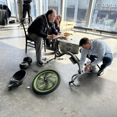 Two people are assembling a Green Machine bike, working together in a well-lit space with large windows. One person is kneeling on the ground, attaching parts of the bike, while the other sits on a chair, reviewing instructions. Bike parts, including the wheel and seat, are spread out on the floor, reflecting a collaborative and charitable team building activity. thumbnail