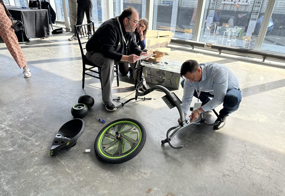 Two people are assembling a Green Machine bike, working together in a well-lit space with large windows. One person is kneeling on the ground, attaching parts of the bike, while the other sits on a chair, reviewing instructions. Bike parts, including the wheel and seat, are spread out on the floor, reflecting a collaborative and charitable team building activity.