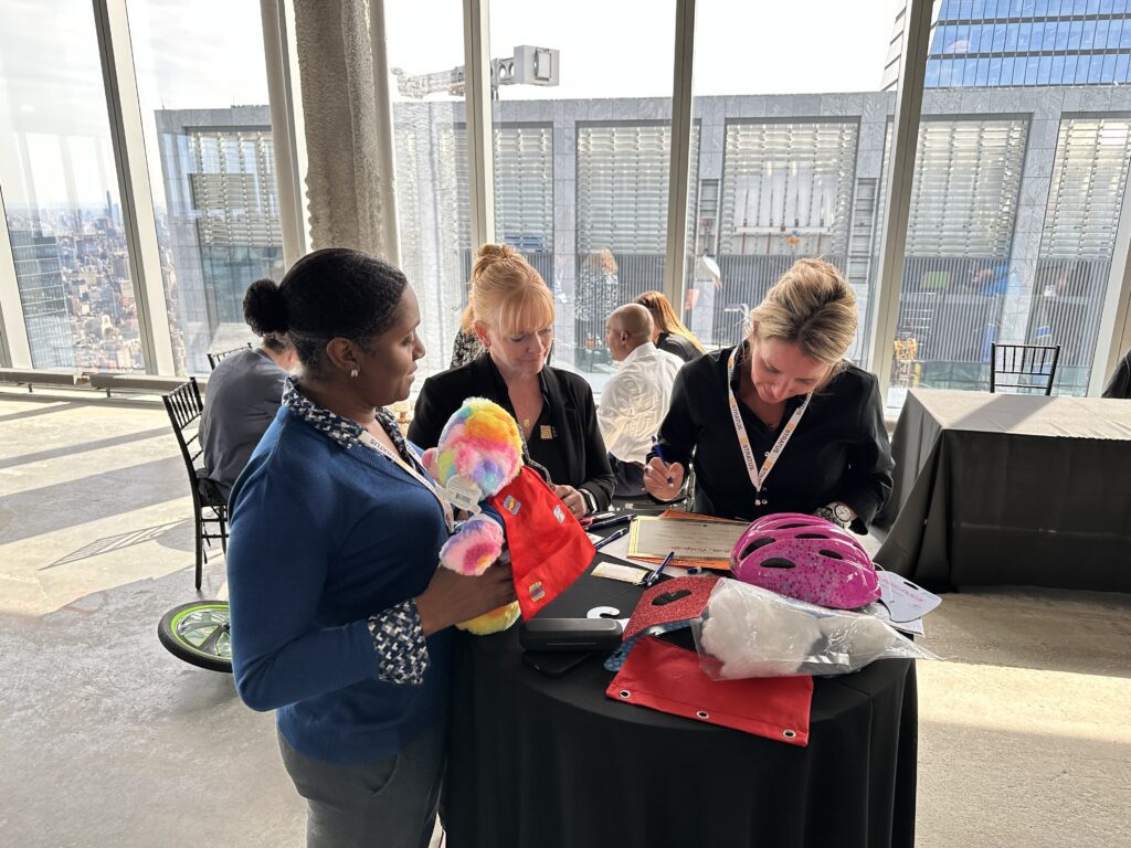 Three individuals at a table working together during a team building activity. One person is holding a colorful teddy bear and superhero-themed cape, while others are focused on paperwork and a helmet on the table.