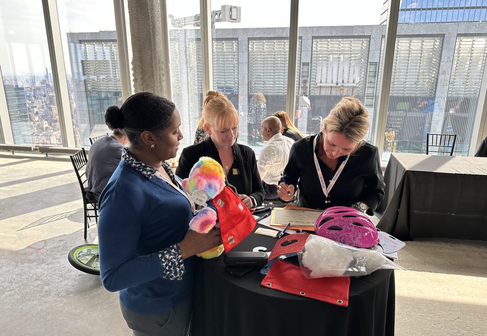 Three individuals at a table working together during a team building activity. One person is holding a colorful teddy bear and superhero-themed cape, while others are focused on paperwork and a helmet on the table.