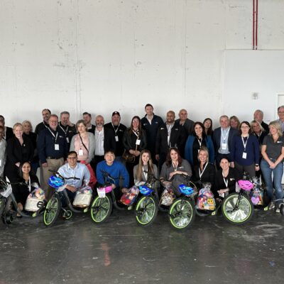 A large group of people poses together indoors, standing behind several Green Machine bikes decorated with colorful baskets and helmets. The group, made up of both seated and standing individuals, is smiling, showing camaraderie and a sense of accomplishment after assembling the bikes during a charitable team building atmosphere. thumbnail