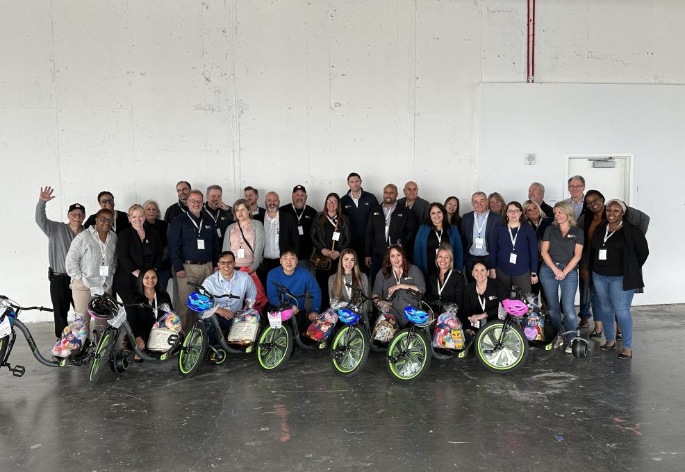 A large group of people poses together indoors, standing behind several Green Machine bikes decorated with colorful baskets and helmets. The group, made up of both seated and standing individuals, is smiling, showing camaraderie and a sense of accomplishment after assembling the bikes during a charitable team building atmosphere.