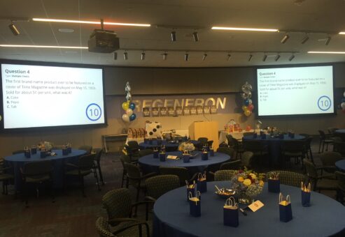 Corporate Quiz Bowl team building event setup with round tables, gift bags, and large screens displaying trivia questions in a well-lit conference room.