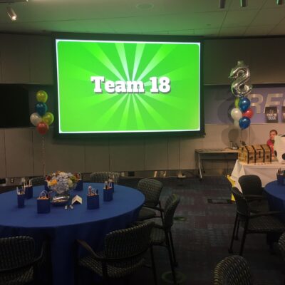 Corporate Quiz Bowl event with a large screen displaying 'Team 18', decorated tables with blue tablecloths, gift bags, balloons, and a playful game atmosphere. thumbnail