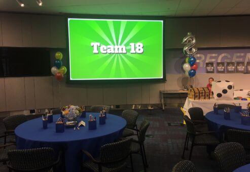 Corporate Quiz Bowl event with a large screen displaying 'Team 18', decorated tables with blue tablecloths, gift bags, balloons, and a playful game atmosphere.