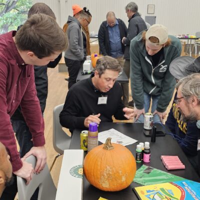 A group of people is gathered around a table, collaborating on what seems to be the design or planning phase of their pumpkin racer project for a team building event. There is a pumpkin on the table, along with various crafting supplies such as glue, markers, and construction paper. The participants appear engaged in a discussion, possibly strategizing or finalizing the design for their pumpkin racer. thumbnail