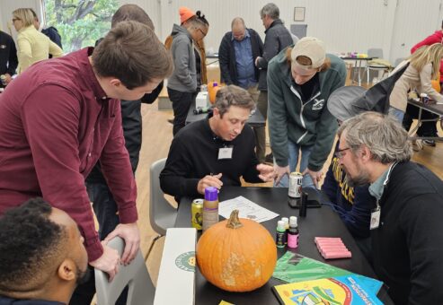 A group of people is gathered around a table, collaborating on what seems to be the design or planning phase of their pumpkin racer project for a team building event. There is a pumpkin on the table, along with various crafting supplies such as glue, markers, and construction paper. The participants appear engaged in a discussion, possibly strategizing or finalizing the design for their pumpkin racer.